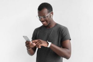 Indoor photo of young African American guy pictured isolated on grey background wearing plastic glasses and wristwatch, typing something on touchscreen of his smartphone with positive relaxed smile