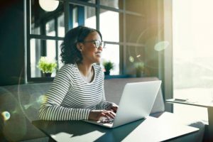 Smiling young African woman wearing glasses looking out of a window while sitting at a table working online with a laptop