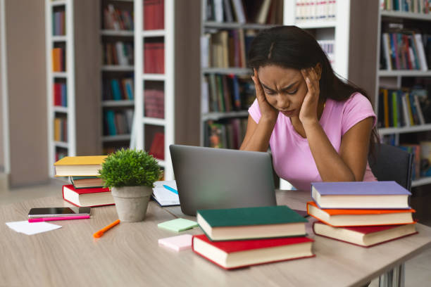 Burnout concept. Tired african american student girl stucked with laptop and books at library, holding her head with both hands, copy space