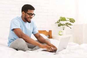 Male programmer typing on laptop in bed, creating program code or website design at home.