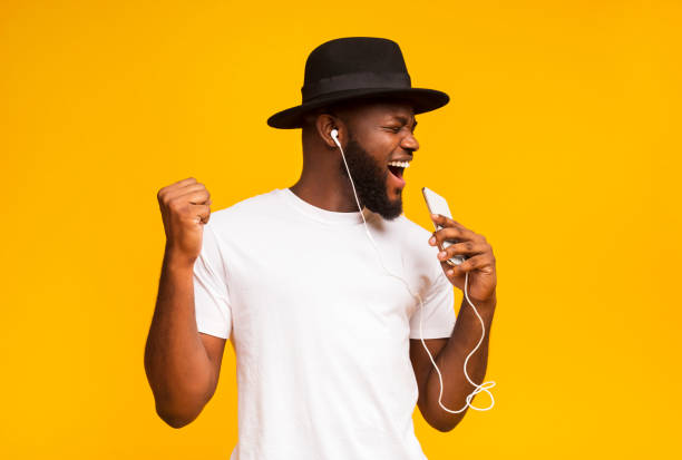 Emotional African guy in black hat singing into smartphone like microphone and listening to music via earphones.