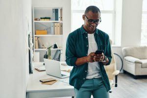 Handsome young African man in shirt using smart phone while standing in the office