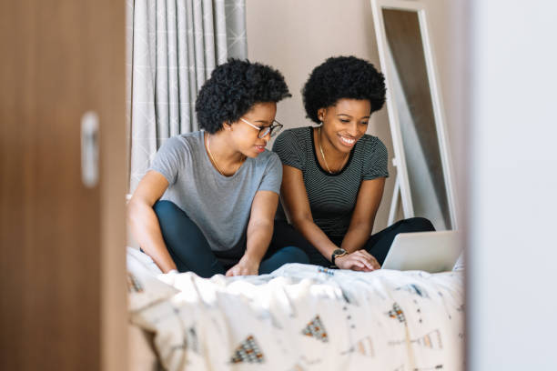 Two black cheerful twin sisters are enjoying their time using a laptop happily at home.