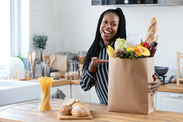 Excited African American woman with grocery paper bag filled with fresh vegetables and bread on kitchen platform looking at camera at home