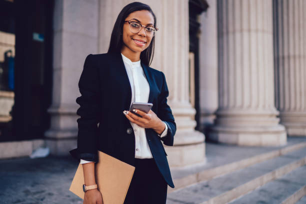 Portrait of successful businesswoman dressed in stylish suit holding in hand folder and mobile phone while standing outdoors near financial office, young woman lawyer using smartphone