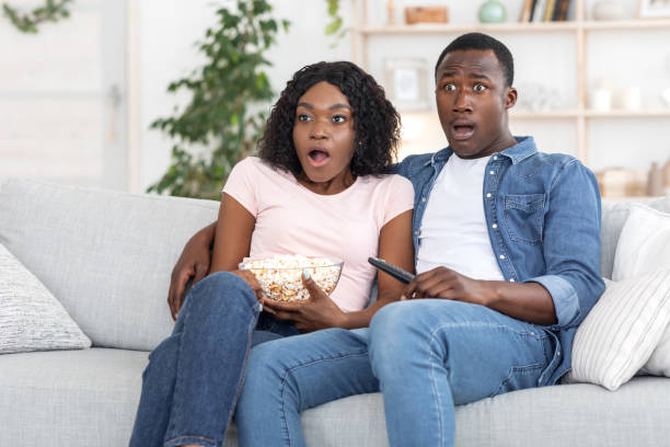 Young man and woman looking in horror, eating popcorn, sitting on sofa at home, as they watch a shocking TV death