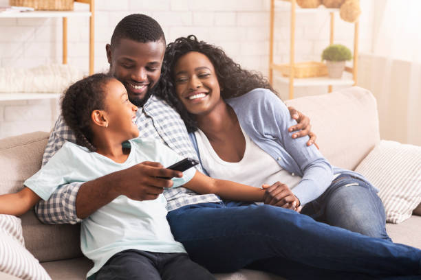 Smiling parents and child relaxing on couch and watching TV at home, man switching channels.