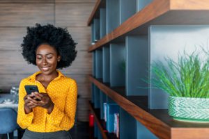 Attractive young African woman holding smart phone and looking at it while standing indoors.