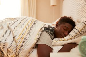 Young African American man lying asleep in bed in the morning, alarm clock in the foreground.