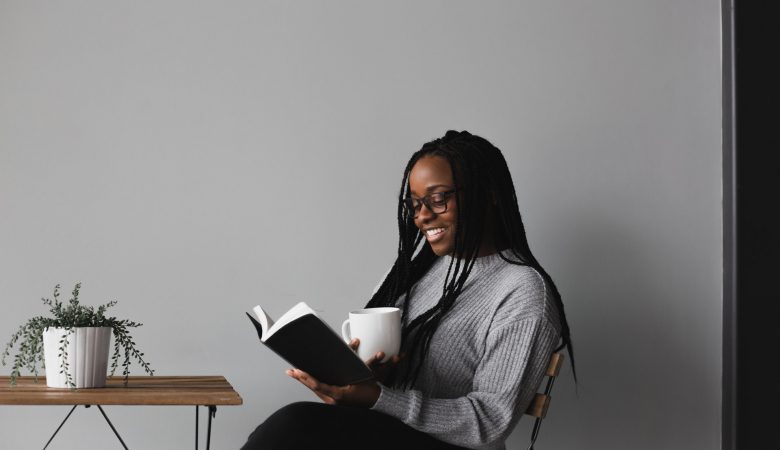 A young lady seated, smiling while reading a book and holding a mug