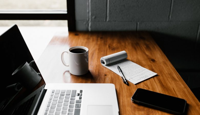 A laptop, cup of coffee, phone, notepad and pen on a desk