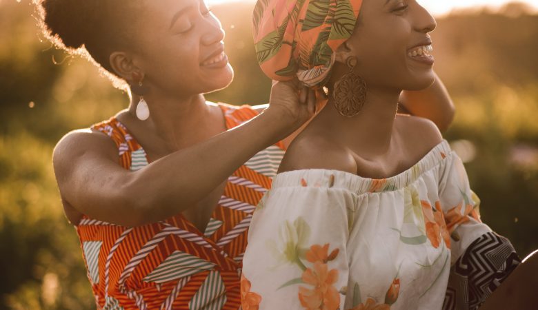 A young woman helping another woman with her headtie. Both smiling.